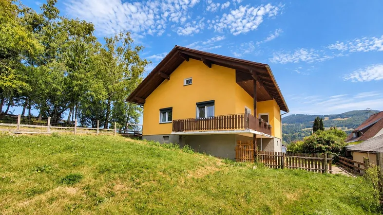 Gelbes Einfamilienhaus auf einem grünen Hügel mit Holzbalkon und Bergblick.