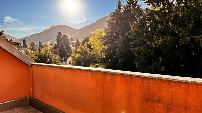 Sonnige Terrasse mit oranger Brüstung und weitem Blick auf die umliegende Berglandschaft und grüne Bäume.