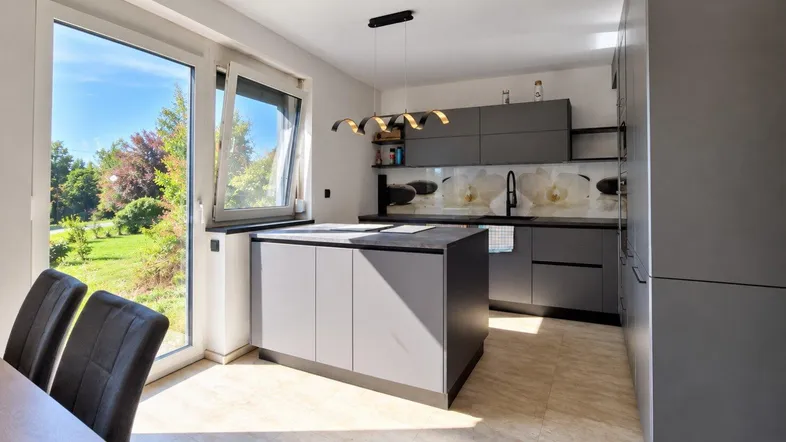 Moderne Küche mit Kücheninsel, grauen Schränken und großem Fenster mit Blick ins Grüne.