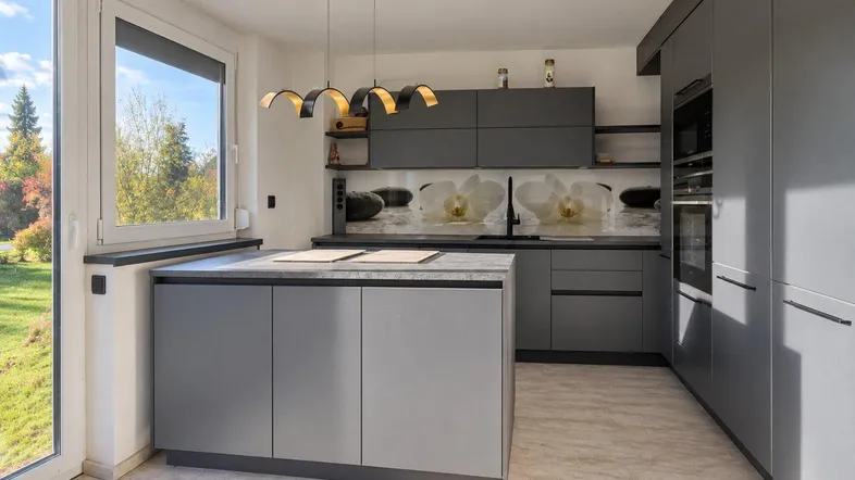 Moderne Küche mit grauen Schränken, Kücheninsel und großem Fenster mit Blick ins Grüne.