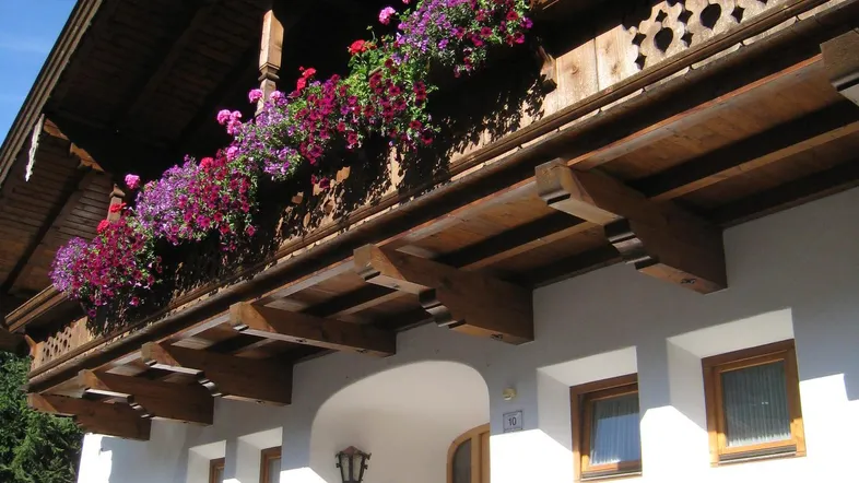 Traditionelles Alpenhaus mit weißer Fassade, holzgeschnitzten Balkonen und bunten Blumenkästen unter blauem Himmel.