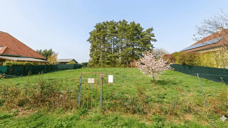 Grünes Baugrundstück mit Rasenfläche, umgeben von Bäumen und Wohnhäusern in ländlicher Umgebung.