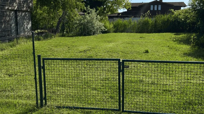 Grünes Grundstück mit Zaun und Tor, umgeben von Bäumen und einem Gebäude im Hintergrund unter blauem Himmel.