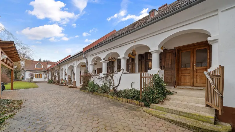 Traditionelles Gebäude mit Arkadengang und gepflastertem Hof, umgeben von Bäumen und Sträuchern.