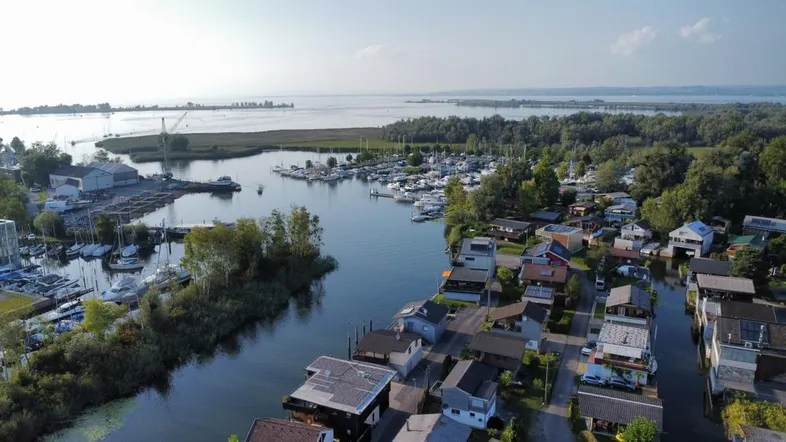 Luftaufnahme einer Siedlung am Seeufer mit Yachthafen und vielen kleinen Häusern, umgeben von grüner Natur.