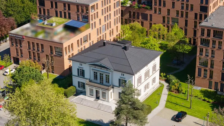Luftaufnahme eines klassischen weißen Gebäudes mit dunklem Dach, umgeben von modernen Backsteingebäuden und viel Grün.