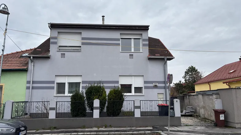 Graue Hausfassade mit weißen Fenstern und Rollläden, umgeben von einem kleinen Vorgarten.
