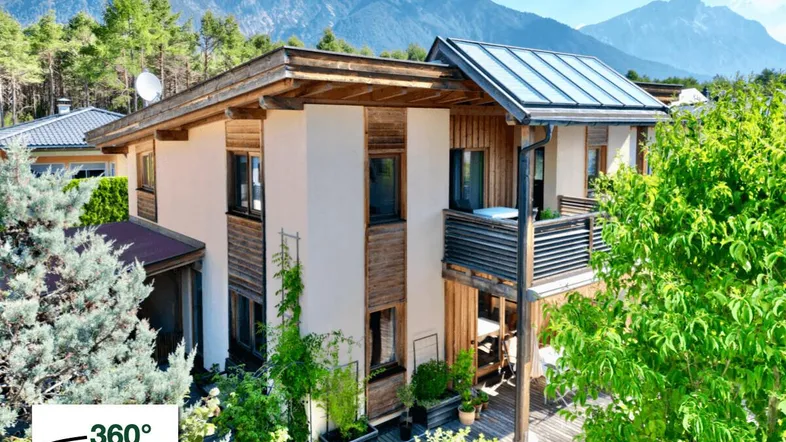 Modernes Haus mit Holzfassade, Balkonen und Solaranlage vor alpiner Bergkulisse.
