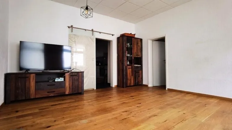Geräumiges Wohnzimmer mit hellem Parkettboden, TV-Möbeln und einem Schiebetürzugang zu einem weiteren Raum.