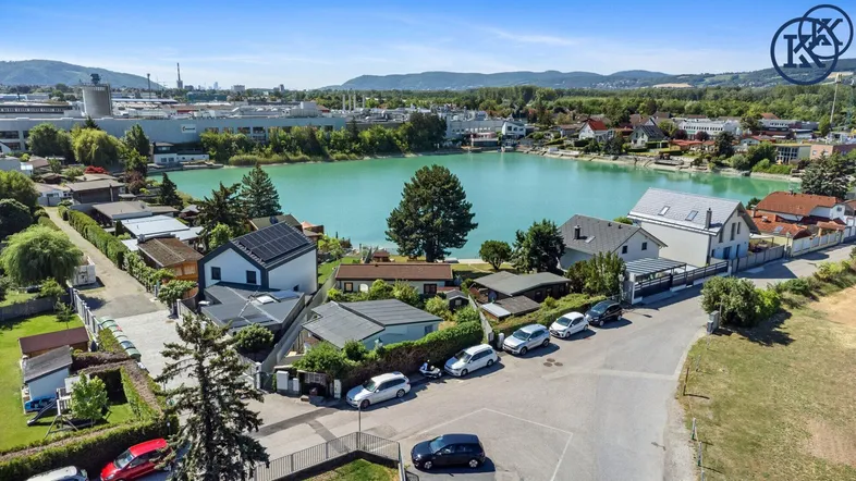 Luftaufnahme einer Wohnsiedlung am Seeufer mit umliegender Landschaft und Bergen im Hintergrund.
