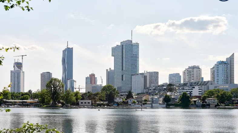 Stadtpanorama mit modernen Hochhäusern und einem See im Vordergrund unter blauem Himmel.