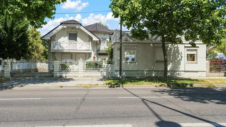 Traditionelles Haus mit weißer Fassade und Balkonen, umgeben von Bäumen an einer Straße.