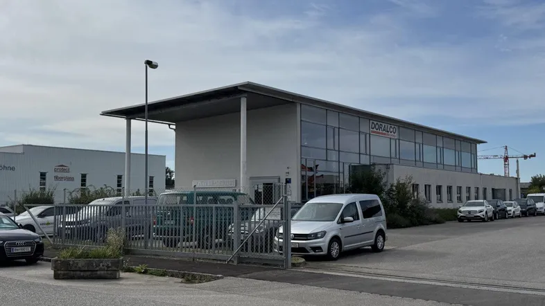 Modernes Geschäftsgebäude mit großen Fenstern und einem Parkplatz davor unter bewölktem Himmel.