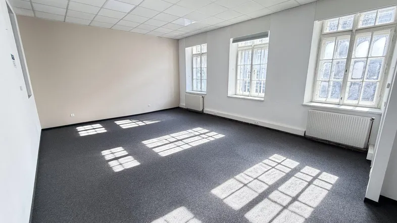 Geräumiges, leeres Büro mit Teppichboden und großen Fenstern, die viel natürliches Licht hereinlassen.