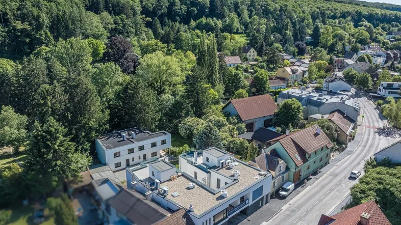 Luftaufnahme eines modernen Gebäudekomplexes mit Flachdächern, umgeben von üppigem Grün und einer Wohnsiedlung.