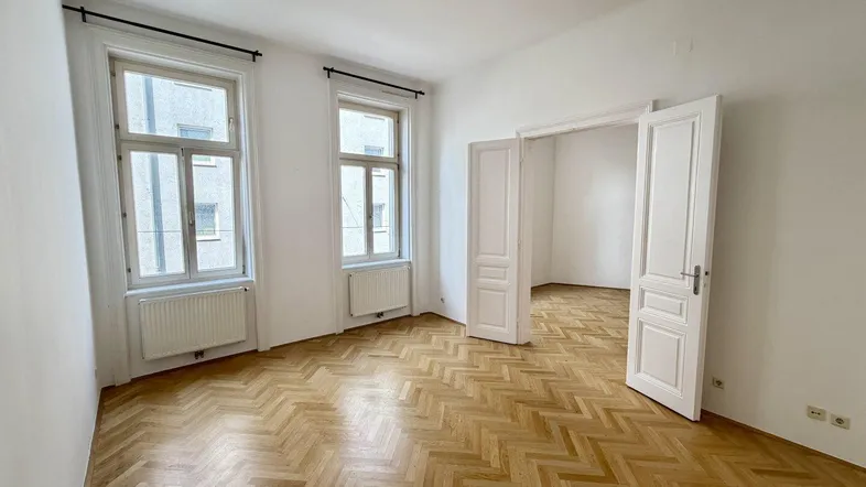 Heller Wohnraum mit Fischgrätparkett, hohen Decken und zwei großen Fenstern, die viel Tageslicht hereinlassen.
