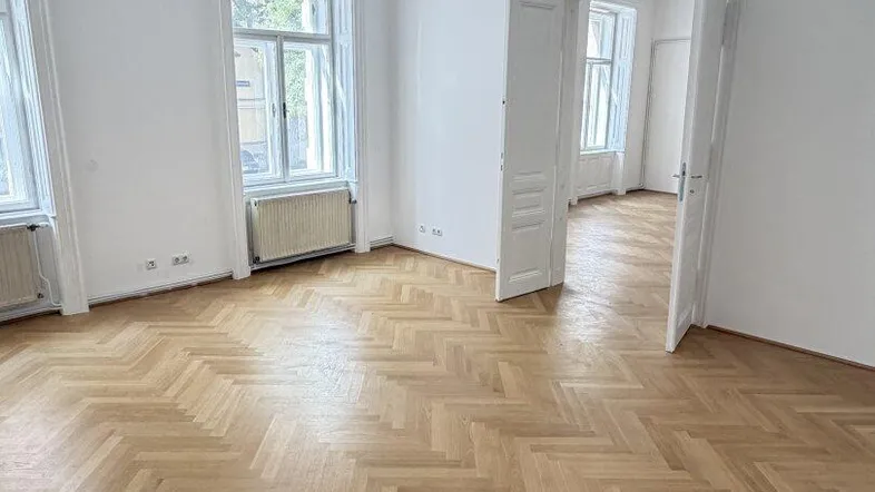 Geräumiges Wohnzimmer mit Fischgrätparkett, hohen Decken und großen Fenstern, die viel Tageslicht hereinlassen.