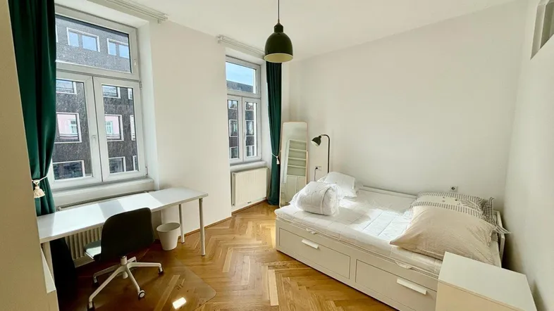 Helles Schlafzimmer mit Parkettboden, Schreibtisch und Blick auf die umliegenden Gebäude.