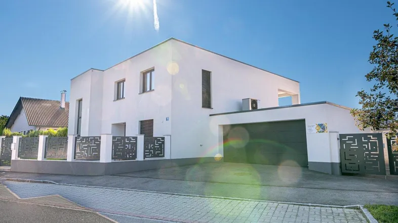 Moderne weiße Villa mit Flachdach, Garage und stilvollem Zaun unter strahlendem Sonnenschein.