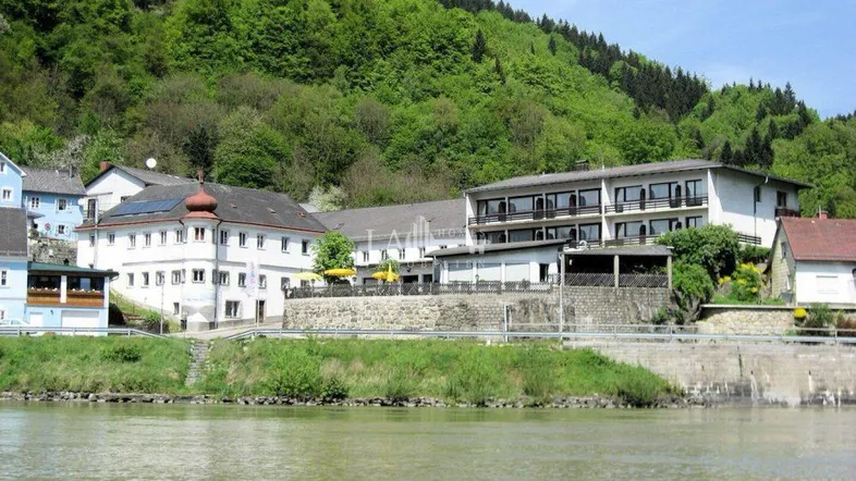 Gebäudekomplex am Flussufer mit grünen Hügeln im Hintergrund und einer Photovoltaikanlage auf dem Dach.