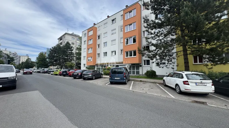 Mehrfamilienhaus mit mehreren Etagen und Parkplätzen entlang der Straße.