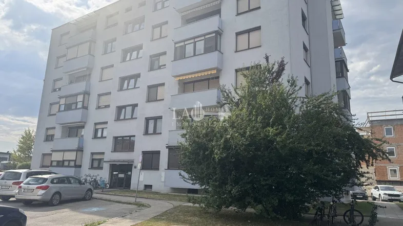 Mehrstöckiges Wohngebäude mit Balkonen, Parkplätzen und Bäumen im Vordergrund.