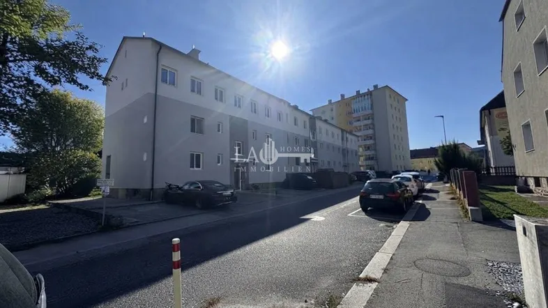 Mehrstöckiges Wohngebäude mit heller Fassade und Parkplätzen an der Straße unter strahlendem Sonnenschein.