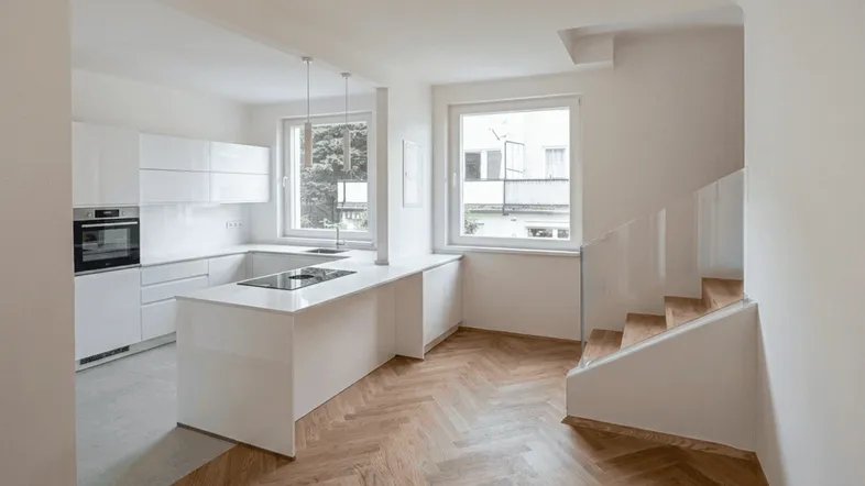 Moderne, offene Küche mit weißer Hochglanzfront, Kochinsel und Fischgrätparkett, daneben eine Treppe mit Glasgeländer.
