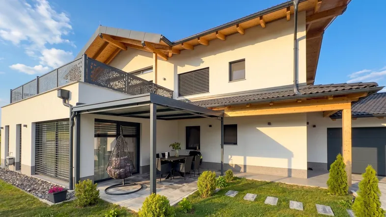 Seitlicher Blick auf das moderne Haus mit überdachter Terrasse und Gartenbereich unter blauem Himmel.