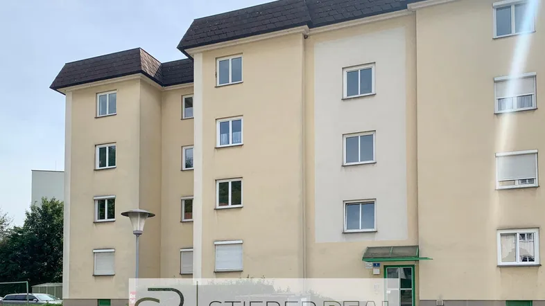 Mehrfamilienhaus mit beiger Fassade und braunem Dach in einer Wohngegend mit Grünflächen.