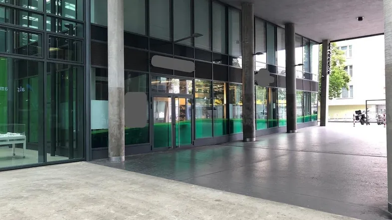 Moderne Gebäudefassade mit großen Glasflächen und einem überdachten Eingangsbereich, der zum Gebäude führt.