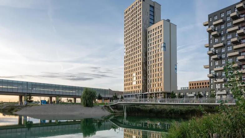 Modernes Hochhaus am Wasser mit Brücke und umliegender Bebauung bei Sonnenuntergang.