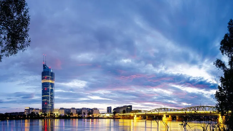 Abenddämmerung über der Donau mit beleuchteter Skyline und Brücke, die sich im Wasser spiegelt.