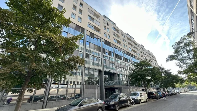 Moderne Bürogebäude-Fassade mit vielen Fenstern, umgeben von Bäumen und parkenden Autos auf der Straße.