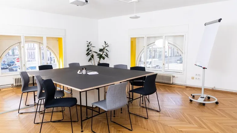 Heller Konferenzraum mit großem Besprechungstisch, Beamer und Whiteboard, umgeben von großen Bogenfenstern.