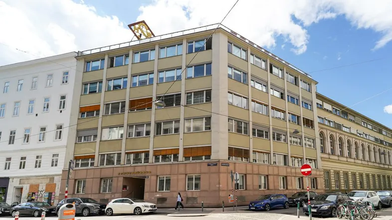 Mehrstöckiges Gebäude mit vielen Fenstern und Autos auf der Straße unter einem bewölkten Himmel.