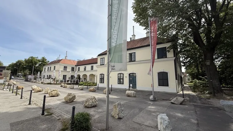 Eine historische Gebäudefassade mit hellen Wänden und dunklen Fenstern, umgeben von Bäumen und einer Straße.