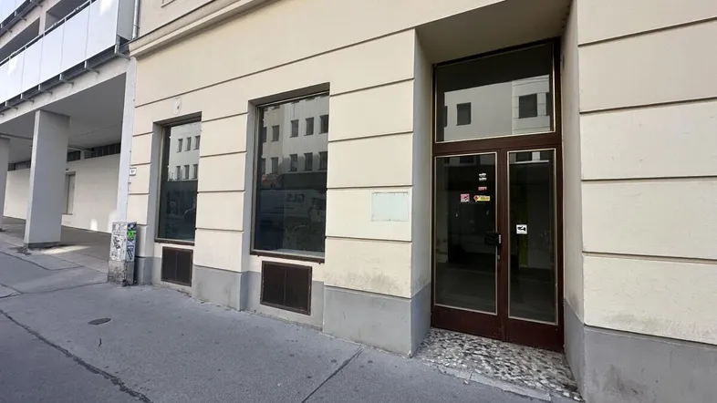 Eingangsbereich eines Geschäftsgebäudes von der Straße aus gesehen mit großen Schaufenstern und beiger Fassade.