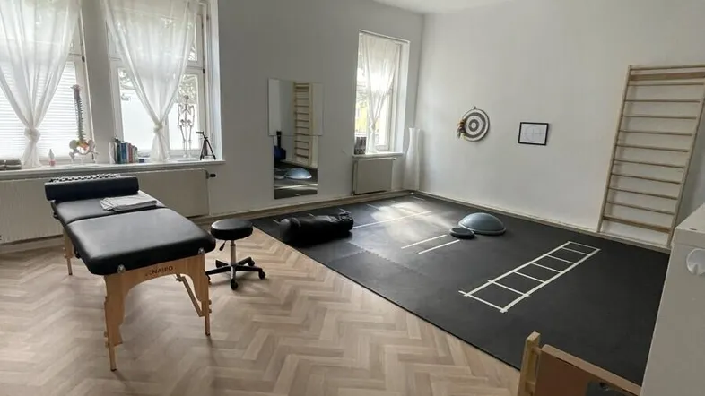 Geräumiger Therapieraum mit Fischgrätparkett, Behandlungsbank und Sportgeräten für physiotherapeutische Anwendungen.