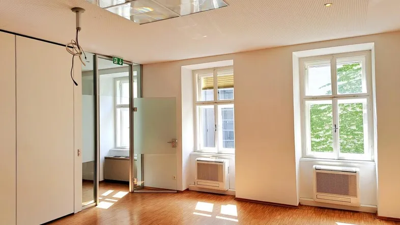 Heller Büroraum mit Holzfußboden, großen Fenstern und moderner Deckenbeleuchtung sowie Glastür.