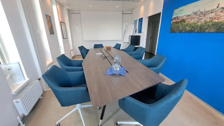 Moderner Konferenzraum mit großem Holztisch, blauen Stühlen und einem Beamer an der Decke.