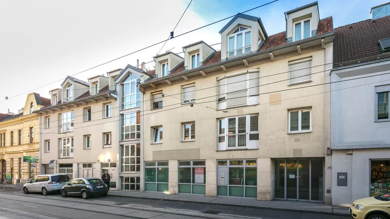 Gebäudeansicht von der Straße mit parkenden Autos und Straßenbahnschienen im Vordergrund.