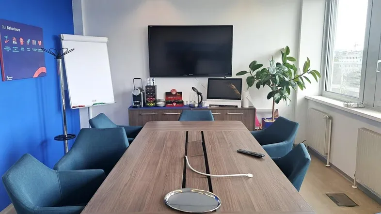 Modern eingerichteter Konferenzraum mit großem Holztisch, blauen Stühlen und einem Wandfernseher.
