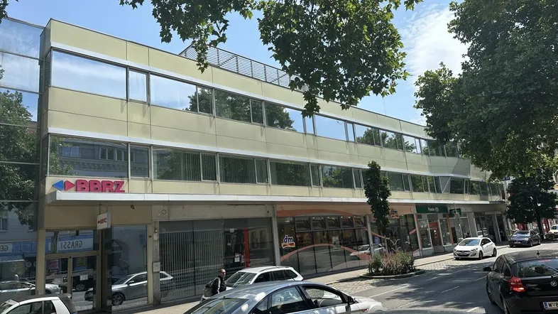 Modernes Geschäftsgebäude mit großen Fenstern, Geschäften im Erdgeschoss und Bäumen entlang der Straße.