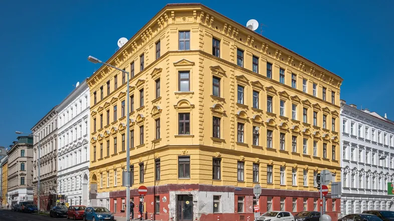 Ein Eckgebäude mit gelber Fassade und vielen Fenstern, umgeben von anderen historischen Gebäuden in einer städtischen Umgebung.
