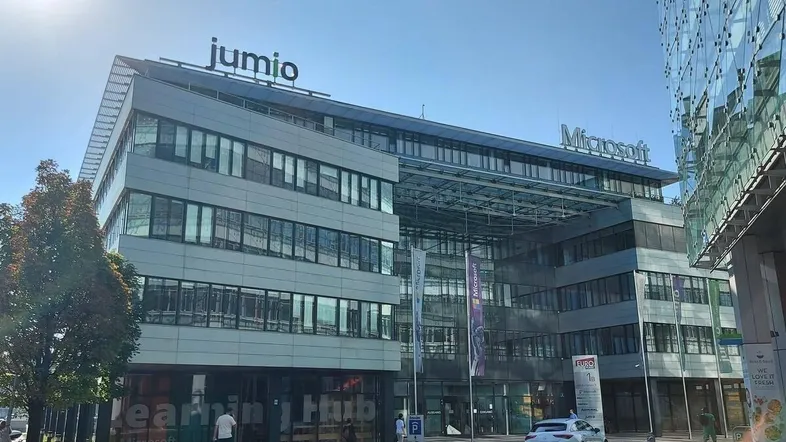 Modernes Bürogebäude mit Glasfassade und Firmenlogos unter blauem Himmel an einem sonnigen Tag.