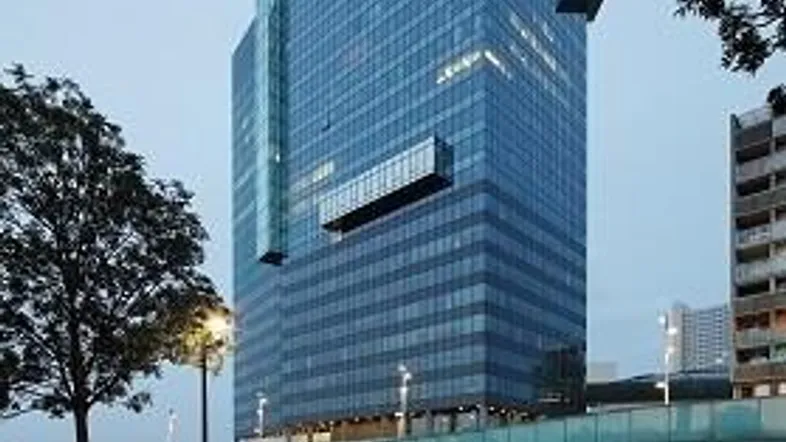 Modernes Hochhaus mit blauer Glasfassade bei Dämmerung, umgeben von Bäumen und einer Straße.