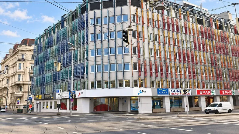 Großes Eckgebäude mit farbiger Fassadenverkleidung und Geschäften im Erdgeschoss an einer belebten Kreuzung.
