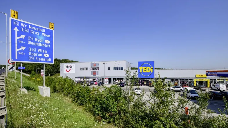 Außenansicht eines Gewerbegebiets mit mehreren Geschäften, Parkplätzen und einer großen blauen Straßentafel.