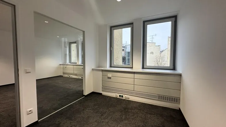 Helles Büro mit dunklem Teppichboden, weißen Wänden und einer modernen Glasabtrennung zu einem weiteren Raum.